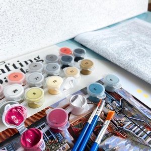 Puede incluir: Un kit de pintura por n&uacute;meros con una variedad de colores de pintura en peque&ntilde;os recipientes, pinceles y un lienzo con una imagen parcialmente pintada de una escena de caf&eacute;. El lienzo tiene la palabra "Cafe" escrita en &eacute;l.