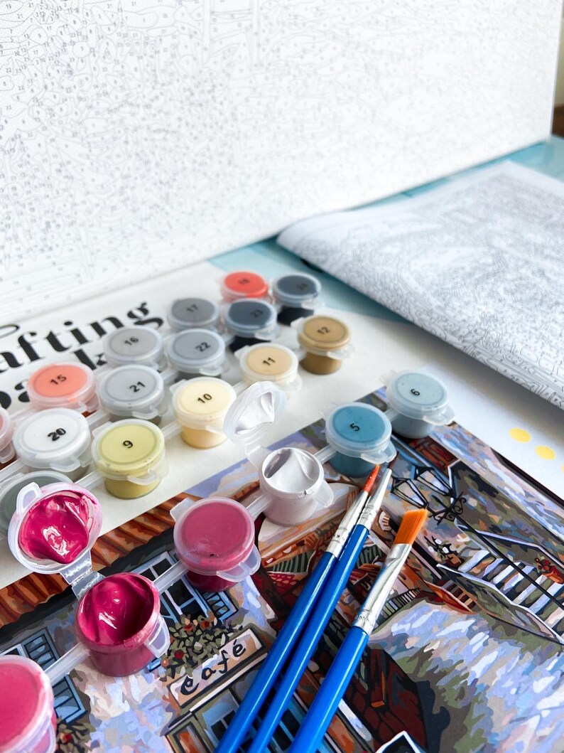 Puede incluir: Un kit de pintura por n&uacute;meros con una variedad de colores de pintura en peque&ntilde;os recipientes, pinceles y un lienzo con una imagen parcialmente pintada de una escena de caf&eacute;. El lienzo tiene la palabra "Cafe" escrita en &eacute;l.