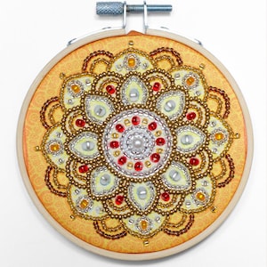 May include: A circular embroidery hoop containing a fabric piece with a beaded mandala design. The pattern features concentric circles of gold, silver, red, and white beads on a yellow fabric background with a subtle orange floral print.