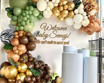 May include: A jungle-themed baby shower balloon arch with green, brown, gold, and white balloons. The arch is decorated with green leaves and a sign that says "Welcome Baby Semaj! We can't wait to meet you!"