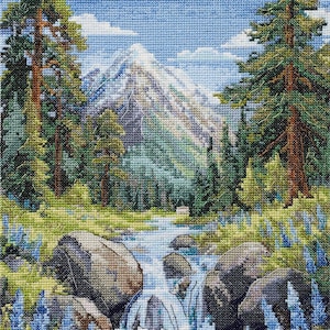 May include: A cross-stitch embroidery depicting a mountain landscape with a snow-capped peak, tall evergreen trees, and a flowing stream. The foreground features large grey rocks surrounded by green grass and patches of blue wildflowers under a blue sky.