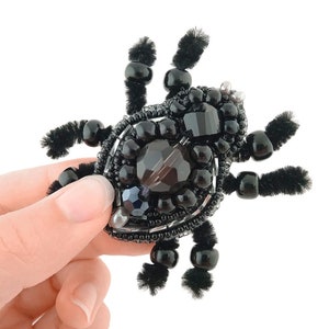 May include: A black beaded spider brooch with a central faceted gem and smaller black beads. The spider's body is adorned with various black beads and gems, and its legs are made of black pipe cleaners.