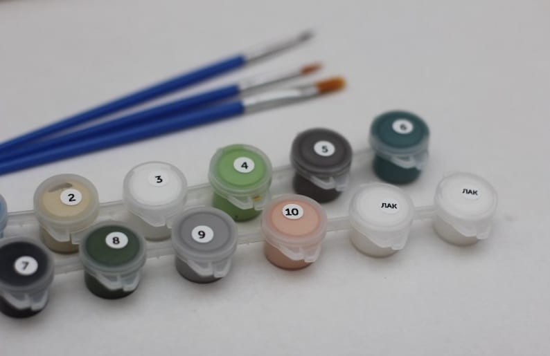 Puede incluir: Un juego de botes de pintura con los n&uacute;meros del 1 al 10, dos de los cuales est&aacute;n etiquetados como 'LAK'. Los botes est&aacute;n dispuestos en una fila sobre una superficie blanca. Hay tres pinceles en el fondo.