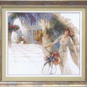May include: A woman in a white dress stands next to a bicycle with a basket of flowers. The bicycle is in front of a white building with a blue door and a vine growing over it.