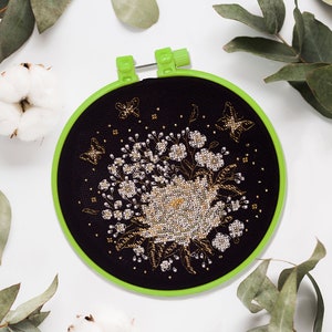 May include: A black embroidery hoop with a green outer ring. The hoop features a floral design with gold and white thread on a black background.