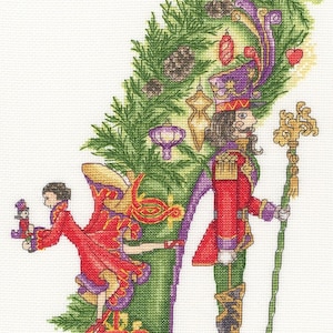 May include: Cross-stitch design featuring a green high-heeled shoe decorated with Christmas ornaments and a fir tree. The shoe holds a Nutcracker soldier and a ballerina in red and purple attire. The design is a festive holiday theme.