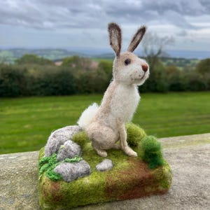 May include: A needle felted rabbit sculpture sitting on a green and brown felt base with rocks and greenery. The rabbit is white with brown ears and a brown nose.