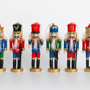 May include: Six colourful wooden nutcracker figurines. Each has a white beard, a gold crown or hat, and a gold base. The nutcrackers are dressed in red, blue, and green uniforms. Some hold staffs or swords. Each is approximately 15 cm tall.