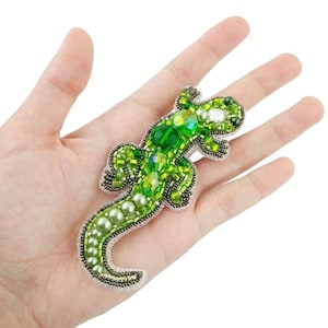 May include: A green beaded lizard brooch with green and white iridescent beads. The brooch is on a hand.
