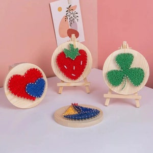 May include: Three wooden string art kits with different designs. The first kit features a red heart with a blue heart inside. The second kit features a red strawberry. The third kit features a green four-leaf clover.