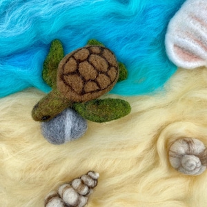 May include: A brown and green felt sea turtle with a brown shell and a gray felt rock. The turtle is sitting on a light yellow felt surface with a blue felt background.