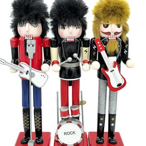 May include: Three wooden nutcracker figures styled as a rock band. The left figure has black hair, a red jacket, and a white guitar. The center figure has black hair, a black jacket, and a drum set labeled ROCK. The right figure has blonde hair, a black jacket, and a red guitar.