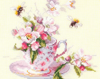 May include: Cross-stitch design of a teacup and saucer filled with pink and white flowers. Two bees fly around the cup. The teacup has a floral pattern and is on a light yellow background.