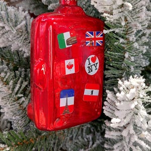 May include: A red suitcase-shaped Christmas ornament decorated with stickers of flags and the phrase "I ❤️ NY." The ornament is suspended by a gold string, set against a frosted Christmas tree.