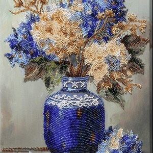 May include: A beaded still life featuring a dark blue vase filled with a bouquet of flowers in shades of blue, cream, and gold. The vase has a detailed white beaded trim. A smaller bouquet sits beside the vase. The artwork is on a neutral background.