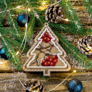 May include: A wooden Christmas tree ornament decorated with red and white beads. The ornament is surrounded by pine branches, pine cones, and white string lights.