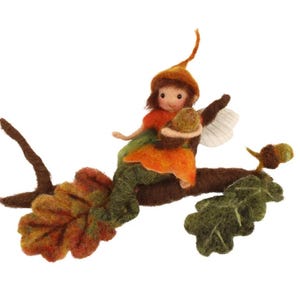 May include: A felt fairy doll with brown hair and a brown acorn hat sits on a branch with green and brown leaves. The fairy is holding a brown acorn in its hand.