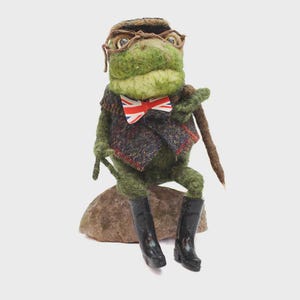 May include: A whimsical felt frog figurine, wearing a tweed vest, Union Jack bow tie, and black boots. The frog is perched on a stone, holding a walking stick, and wearing glasses and a hat. The frog is green and brown.