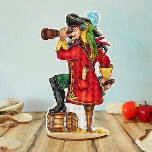 May include: A cross-stitch figurine of a pirate captain with a parrot on his shoulder, holding a spyglass and a rolled-up map. He is standing on a wooden barrel and wearing a red coat, green pants, and a black hat.