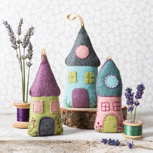 May include: Three felt house decorations in pastel shades of green, blue, and pink, each with a contrasting roof. The houses have windows, doors, and hanging loops. Lavender sprigs and thread spools are also visible, adding to the handcrafted aesthetic.