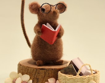 May include: A brown felt mouse figurine wearing small round glasses, holding a miniature red book. The mouse is standing on a wooden slice, with a small basket of books and flowers nearby. The mouse has a long brown tail.