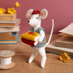 May include: A white felt mouse figurine wearing a blue vest with a red bow on its head. The mouse is holding two stacked felt books, one yellow with a red flower design and one red. The mouse has a long tail and stands upright.
