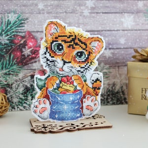 May include: A cross-stitch of a cute tiger cub holding a blue bag with a red bow. The tiger is wearing a green scarf and has orange and black stripes. The tiger is standing on a wooden base with a snowflake design.