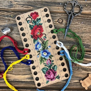 May include: A wooden embroidery floss organizer with 26 holes. The organizer has a floral cross-stitch design with red, blue, and pink flowers. There are strands of floss in red, blue, green, and white.