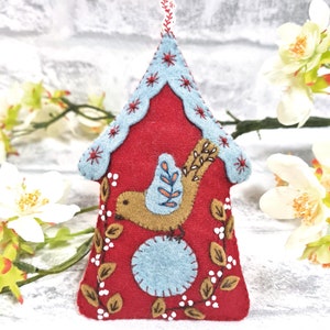 May include: A felt birdhouse ornament with a red exterior, blue roof, and a brown bird with blue wings. The birdhouse is decorated with white and brown stitching.