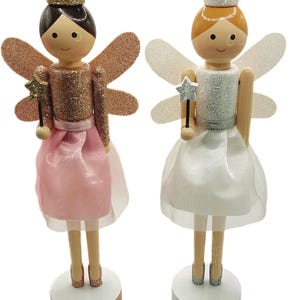 May include: Two wooden fairy figurines. One fairy has brown hair, a gold crown, and a pink dress. The other fairy has blonde hair, a silver crown, and a white dress. Both fairies have glittery wings and hold star-shaped wands.