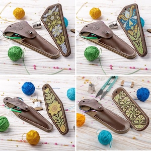 May include: A collection of brown leather needle cases with decorative cross-stitch designs. Each case is open, revealing sewing tools. Colorful yarn balls and sewing accessories are scattered around the cases on a light wood surface.