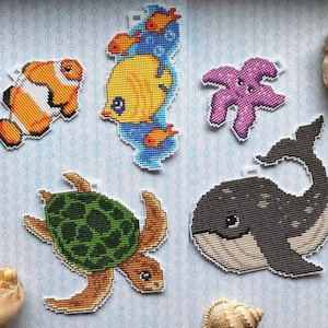 May include: Five colorful cross-stitch ornaments featuring a whale, turtle, starfish, clownfish, and a yellow fish with coral and bubbles. The ornaments are on a blue and white patterned background.