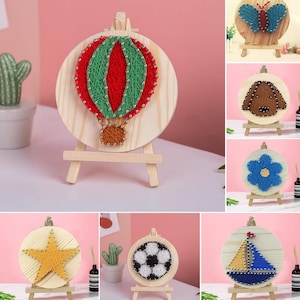 May include: A collection of handmade string art on small wooden easels. The designs include a hot air balloon, butterfly, dog, flower, star, soccer ball, and sailboat. Each piece features colorful thread and small silver pins on a round wooden base.