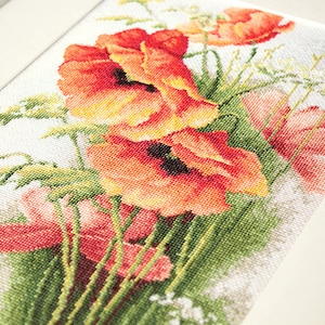 May include: Cross-stitch artwork featuring a vibrant bouquet of red and orange poppies with green foliage. The needlework is framed in a white border, showcasing the detailed floral design. The piece is a close-up view.
