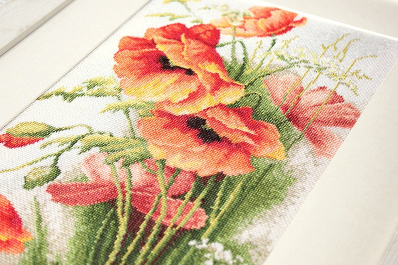 May include: Cross-stitch artwork featuring a vibrant bouquet of red and orange poppies with green foliage. The needlework is framed in a white border, showcasing the detailed floral design. The piece is a close-up view.