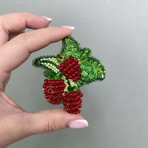 May include: A beaded brooch featuring a cluster of red berries with green leaves. The berries are made of small red beads and the leaves are made of green beads.