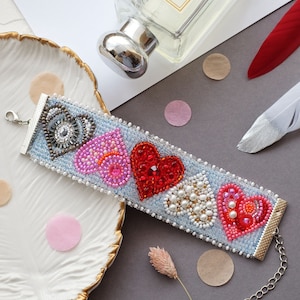 May include: A beaded bracelet with five heart designs in shades of red, pink, and white, set against a light blue background. The bracelet is adorned with beads and pearls, and has a silver clasp. A perfume bottle and decorative feathers are also in the image.