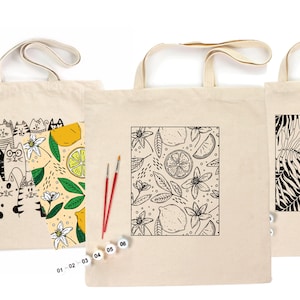 May include: Three canvas tote bags with different designs. One features a cat illustration, another has a lemon and floral pattern, and the third displays a black and white tropical leaf design. Paintbrushes and paint pots are included.
