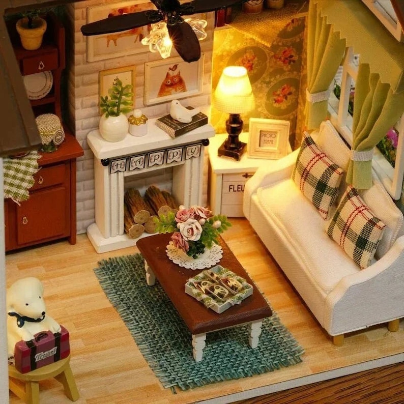 Puede incluir: Una sala de estar de casa de mu&ntilde;ecas en miniatura con un sof&aacute; blanco, una chimenea, una mesa de caf&eacute;, una alfombra y una peque&ntilde;a figura de perro. La habitaci&oacute;n est&aacute; decorada con un arreglo floral, una l&aacute;mpara y cuadros enmarcados.