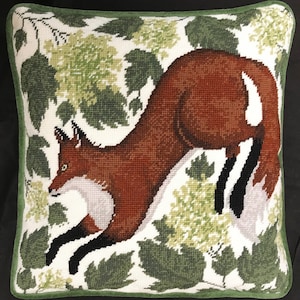 May include: A red fox jumps through a green leafy background. The fox has a white chest and a bushy tail.