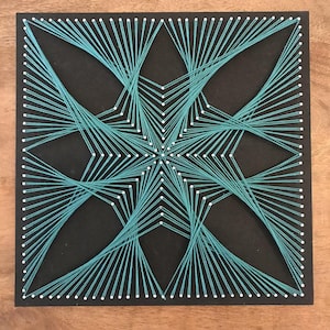May include: A square black board featuring a geometric string art design created with teal thread wrapped around white nails. The threads are arranged in a symmetrical star-like pattern, radiating from the centre towards the edges of the board.