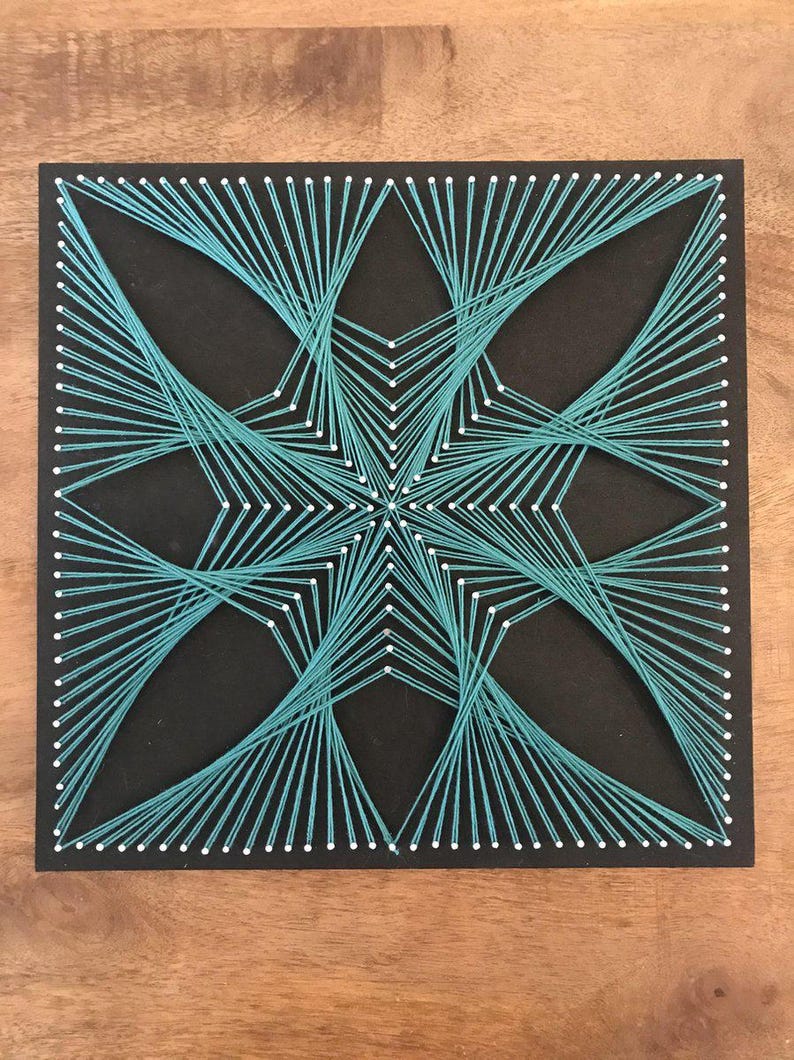 May include: A square black board featuring a geometric string art design created with teal thread wrapped around white nails. The threads are arranged in a symmetrical star-like pattern, radiating from the centre towards the edges of the board.
