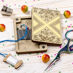 May include: A wooden needle case with a decorative geometric design and a magnetic closure. The case is open, revealing needles and blue thread. A thimble, scissors, and a spool of thread are also present, along with colorful beads and small fruit.