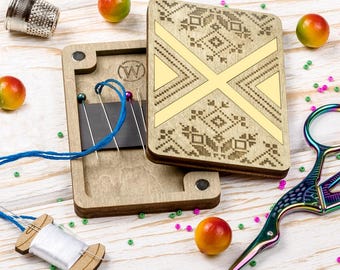 May include: A wooden needle case with a decorative geometric design and a magnetic closure. The case is open, revealing needles and blue thread. A thimble, scissors, and a spool of thread are also present, along with colorful beads and small fruit.