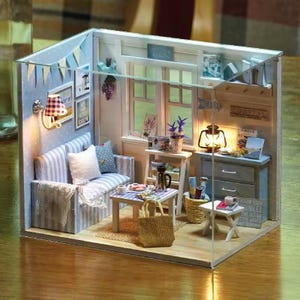 May include: A miniature dollhouse interior with a coastal theme. The scene includes a striped sofa, small tables, a lamp, and decorative items. The walls are painted in light blue and white, with framed artwork and a banner. A small tote bag sits on the floor.