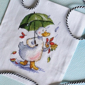 May include: Cross-stitch artwork featuring a cartoon duck under a green umbrella, holding autumn leaves. The duck is walking in the rain, with falling leaves and raindrops. The artwork is framed by a black and white rope.