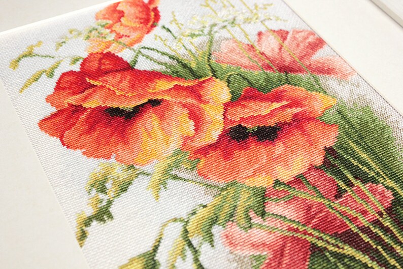 May include: Cross-stitch artwork featuring a vibrant bouquet of red and orange poppies with green foliage. The needlework is detailed, showcasing the texture of the stitches. The artwork is framed in a white border.