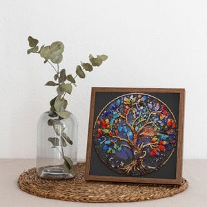 May include: A framed circular artwork depicting a beaded tree with vibrant leaves and a brown trunk. The artwork is set against a dark background and framed in a wooden frame. A clear glass vase with green leaves is to the left.