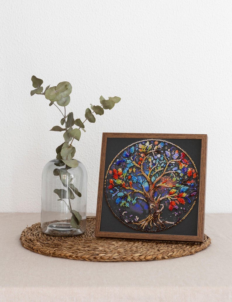 May include: A framed circular artwork depicting a beaded tree with vibrant leaves and a brown trunk. The artwork is set against a dark background and framed in a wooden frame. A clear glass vase with green leaves is to the left.