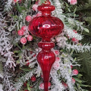 May include: A vibrant red Christmas ornament with a textured, bulbous design. The ornament has a long, pointed bottom and is suspended by a gold string. The background features frosted greenery and red berries, creating a festive holiday scene.